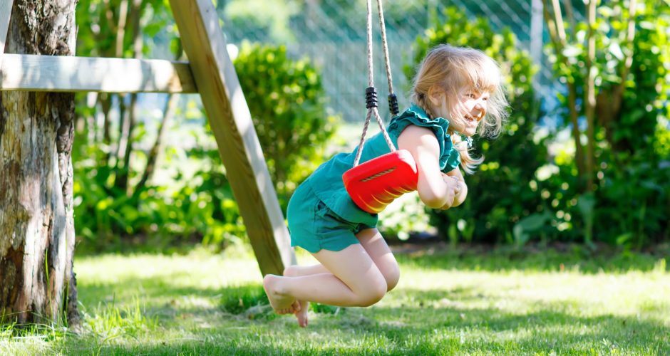 Happy little preschool girl having fun on swing in domestic garden. Healthy toddler child swinging on sunny summer day. Children activity outdoor, active smiling kid laughing.